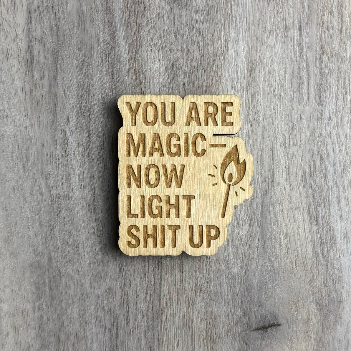Wooden magnet engraved with the bold, all-caps phrase "YOU ARE MAGIC — NOW LIGHT SHIT UP." To the right of the text is an engraved illustration of a lit match with stylized flame lines. The magnet has a jagged, organic outline and is displayed on a gray-toned wood background.