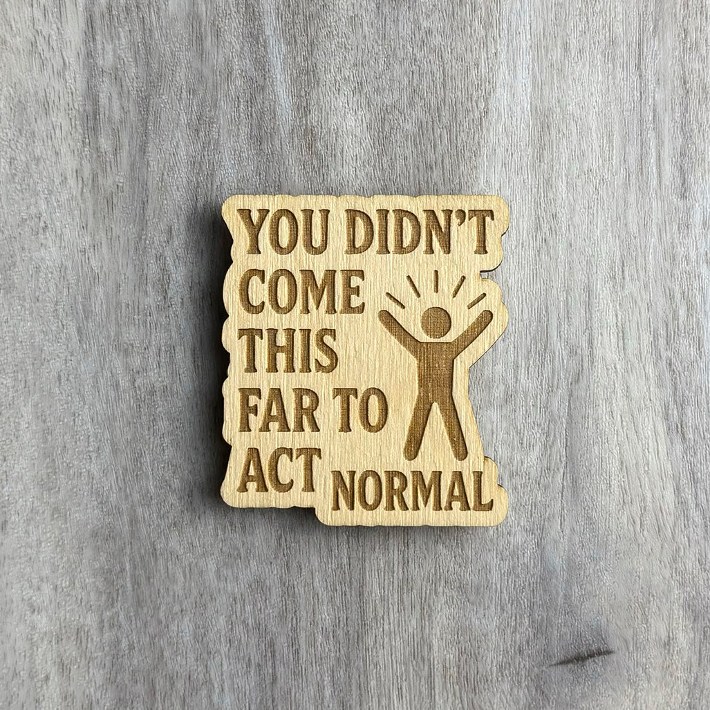 Wooden magnet engraved with the phrase "YOU DIDN’T COME THIS FAR TO ACT NORMAL" in all caps. The right side features a figure with arms raised and radiating lines above its head, symbolizing energy or celebration. The magnet has an irregular, wavy border and is placed on a light gray wood surface.