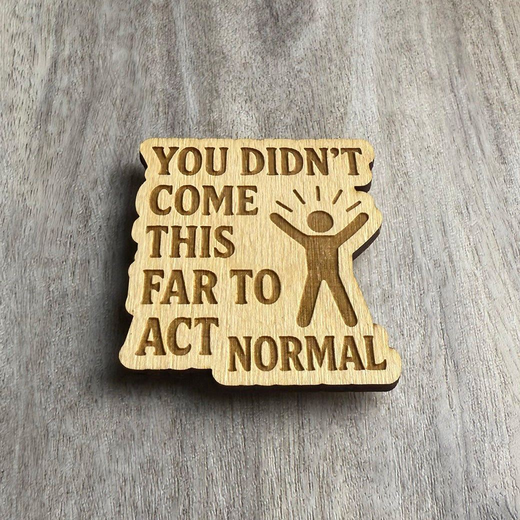 Angled view of a wooden magnet engraved with the bold text "YOU DIDN’T COME THIS FAR TO ACT NORMAL" alongside a stick-figure style person with arms raised and lines indicating excitement. The wood piece has soft, uneven edges and rests on a textured wood background.