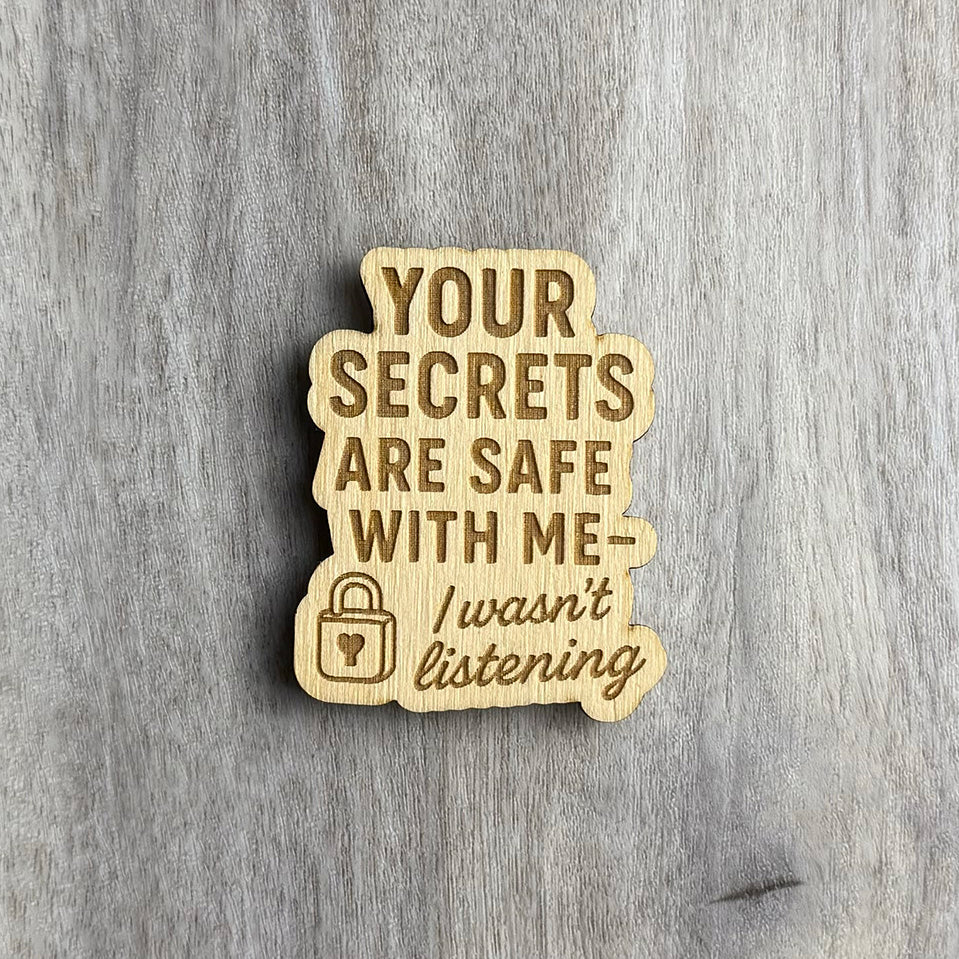 Wooden magnet engraved with the phrase "YOUR SECRETS ARE SAFE WITH ME — I wasn't listening" in a mix of bold block and cursive fonts. A small lock icon with a heart-shaped keyhole is engraved on the lower left. The magnet has a soft, bubbly border and is placed on a light gray wood background.