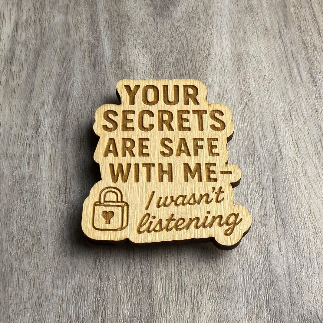 Angled view of a wooden magnet engraved with the text "YOUR SECRETS ARE SAFE WITH ME — I wasn't listening," combining uppercase block letters with cursive script. A heart-lock icon sits to the left of the lower text. The magnet is shaped with rounded, organic curves and rests on a textured wood grain surface.