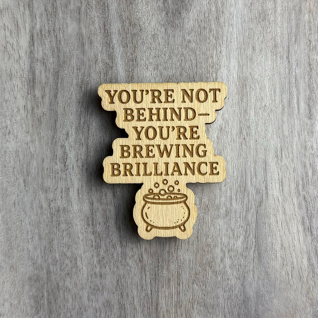 Wooden magnet engraved with the uplifting phrase "YOU'RE NOT BEHIND — YOU'RE BREWING BRILLIANCE" in uppercase letters. Below the text is a bubbling cauldron graphic, symbolizing creativity in progress. The magnet has a tiered, stacked shape and sits on a light gray wood surface.