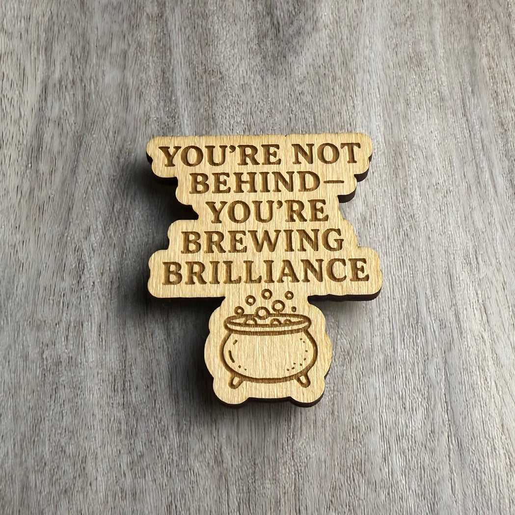 Angled view of a wooden magnet featuring the phrase "YOU'RE NOT BEHIND — YOU'RE BREWING BRILLIANCE" with an engraved bubbling cauldron below the text. The magnet is shaped like stacked blocks of text with a rounded bottom and is displayed on a wood-textured background.