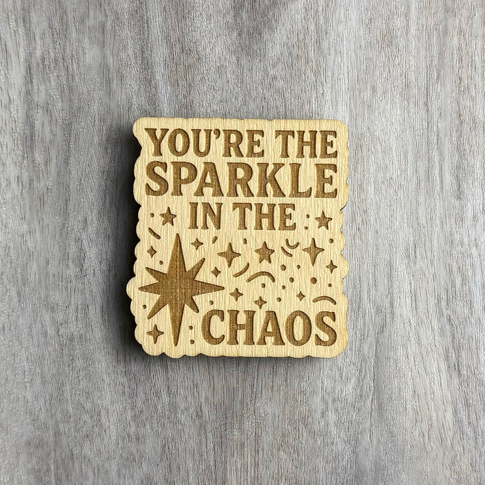 Wooden magnet engraved with the phrase "YOU’RE THE SPARKLE IN THE CHAOS" in uppercase letters. The background is filled with engraved stars, sparkles, dots, and swirls, including a large starburst shape on the left. The magnet has a scalloped border and is placed on a light gray wood surface.