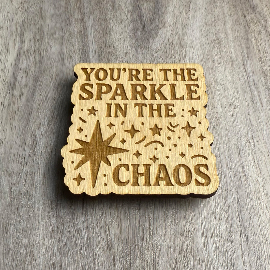 Angled view of a wood magnet featuring the engraved phrase "YOU’RE THE SPARKLE IN THE CHAOS" surrounded by scattered stars, confetti shapes, and a large starburst. The cutout has wavy, rounded edges and rests on a wood-textured background.