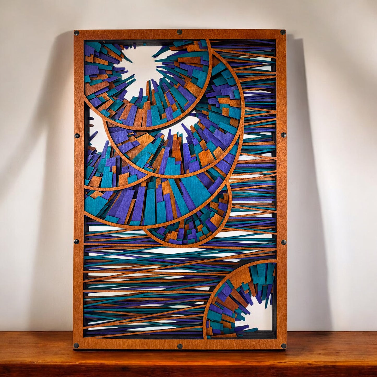 Wood wall art displayed above a wooden surface, centered and well-lit, showing crisp outlines of nested spirals in a burst-like pattern with horizontal color-threaded accents.