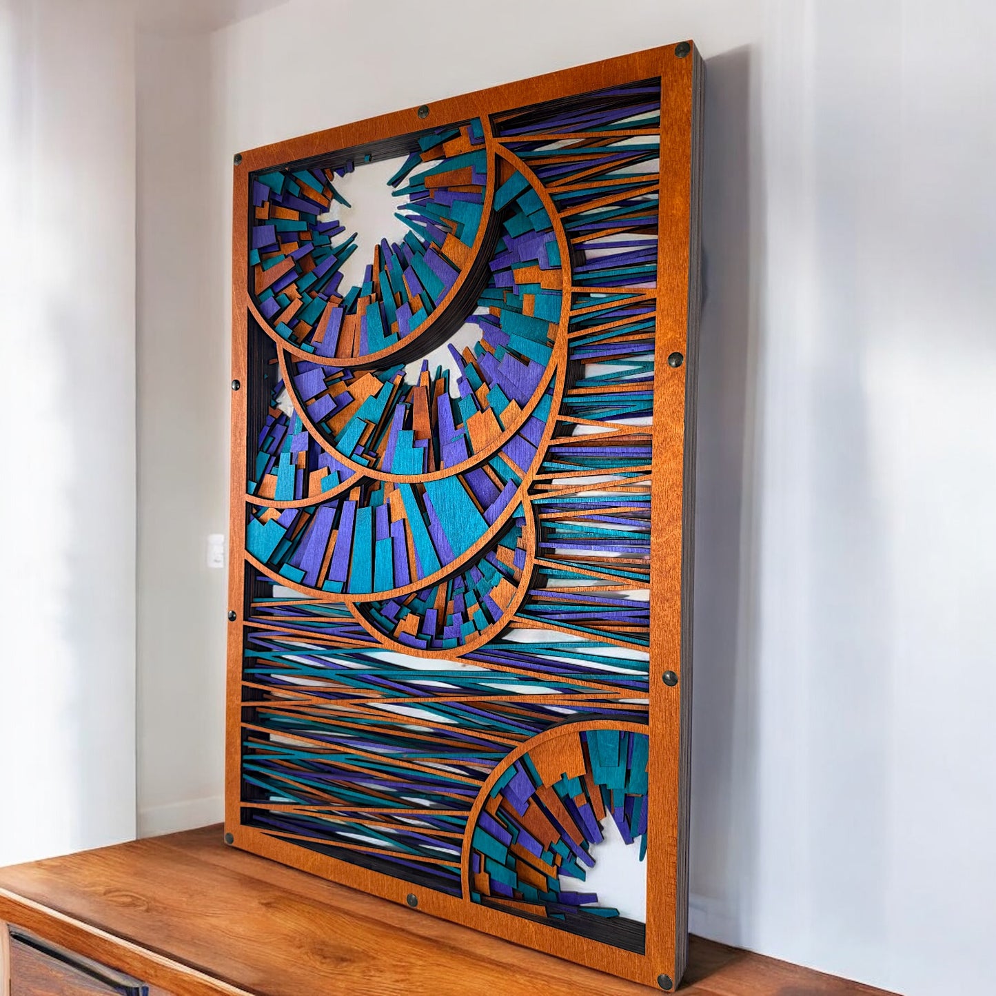 Angled shot of the wall art leaning on a wooden cabinet, showcasing the warm stained frame and the colorful layers creating a dynamic, optical energy effect.