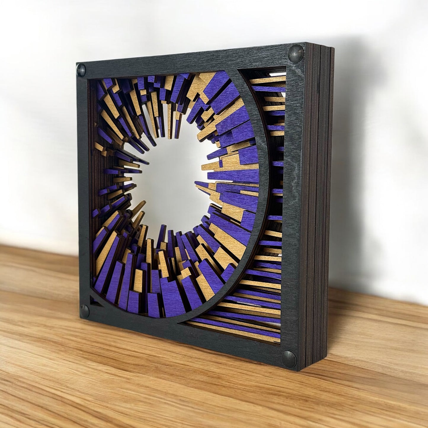 Metrorhythm V1 placed on a wood surface, catching light across its dimensional birch layers in black, purple, and gold.