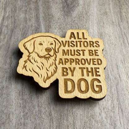 Angled view of a laser-cut wooden magnet with an engraved golden retriever and the text 'ALL VISITORS MUST BE APPROVED BY THE DOG', showing the depth of the cut and thickness of the wood, placed on a gray wood surface.
