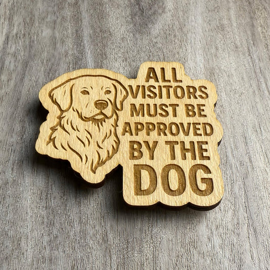 Angled view of a laser-cut wooden magnet with an engraved golden retriever and the text 'ALL VISITORS MUST BE APPROVED BY THE DOG', showing the depth of the cut and thickness of the wood, placed on a gray wood surface.