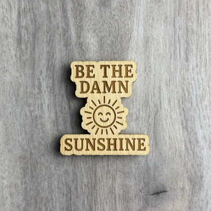 Laser-cut wooden magnet with the phrase Be the Damn Sunshine engraved in bold lettering, separated by a smiling sun illustration in the center, placed on a gray wood background.