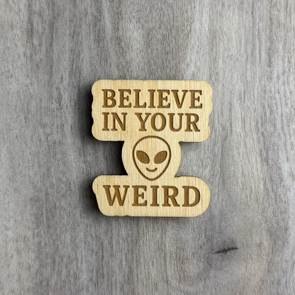 Laser-cut wooden magnet engraved with the phrase Believe in Your Weird, featuring a stylized alien face in the center between the text, displayed on a gray wood background.