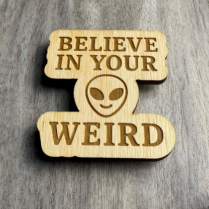 Angled view of a laser-cut wooden magnet engraved with the phrase Believe in Your Weird, featuring a bold alien face in the center, highlighting the depth of the engraving and the layered wood construction on a gray wood surface.
