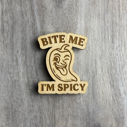 Laser-cut wooden magnet engraved with the phrase Bite Me I'm Spicy, featuring a playful chili pepper illustration with a winking face and tongue sticking out, placed on a gray wood background.