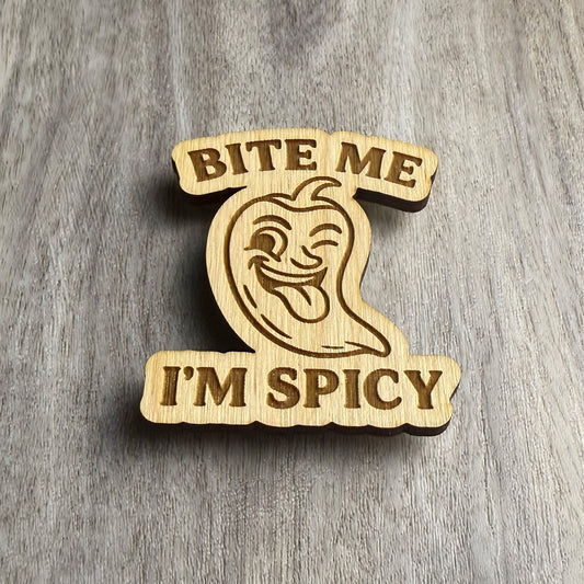 Angled view of a laser-cut wooden magnet showing the phrase Bite Me I'm Spicy and a cheeky chili pepper face in the center, highlighting the depth and contour of the engraved wood on a gray surface.