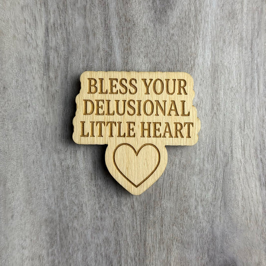 Laser-cut wooden magnet with the phrase Bless Your Delusional Little Heart engraved in bold text above a simple outlined heart shape, set against a gray wood background.