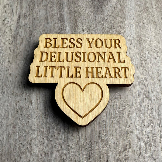 Angled view of a laser-cut wooden magnet engraved with Bless Your Delusional Little Heart and a heart icon below the text, showing the depth of the engraving and wood thickness on a gray surface.