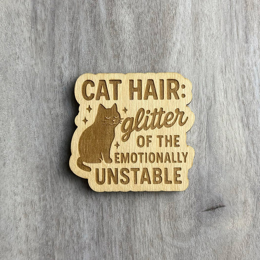 Laser-cut wooden magnet engraved with the phrase Cat Hair: Glitter of the Emotionally Unstable, featuring a smug-looking cat illustration surrounded by sparkles, placed on a gray wood background.