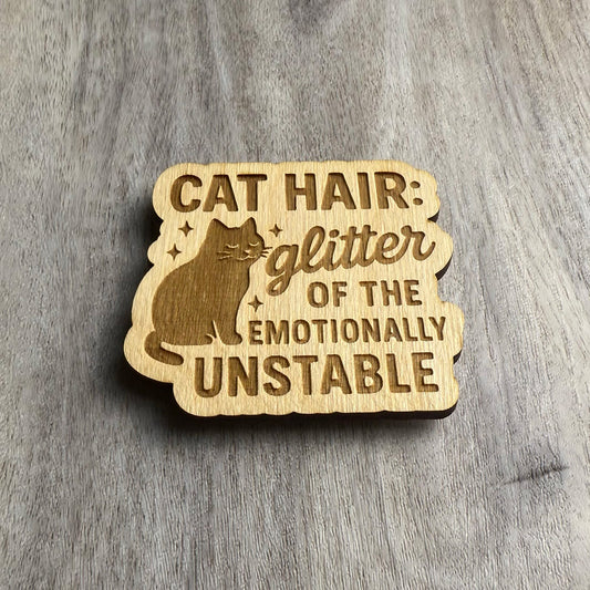 Angled view of a laser-cut wooden magnet with the engraved phrase Cat Hair: Glitter of the Emotionally Unstable and a cat with sparkles, showing the engraved depth and layered wood on a gray surface.