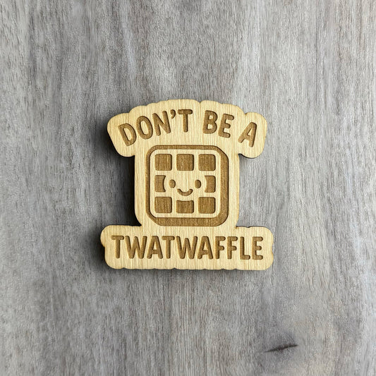 Laser-cut wooden magnet engraved with the phrase Don’t Be a Twatwaffle, featuring a smiling waffle character in the center, all set against a gray wood background.