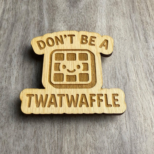 Angled view of a laser-cut wooden magnet with the engraved phrase Don’t Be a Twatwaffle and a happy waffle illustration in the middle, showing depth and wood layering on a gray surface.