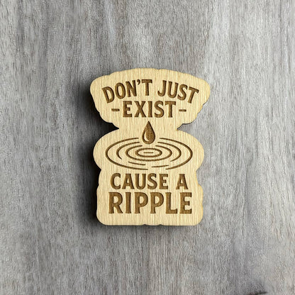 Laser-cut wooden magnet engraved with the phrase Don’t Just Exist — Cause a Ripple, featuring a water droplet above concentric ripple rings in the center, set on a gray wood background.