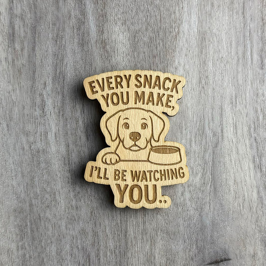 Angled view of a laser-cut wooden magnet engraved with the phrase Every Snack You Make, I’ll Be Watching You, featuring a cute dog illustration next to a food bowl, showing engraving depth and wood layering on a gray wood surface.