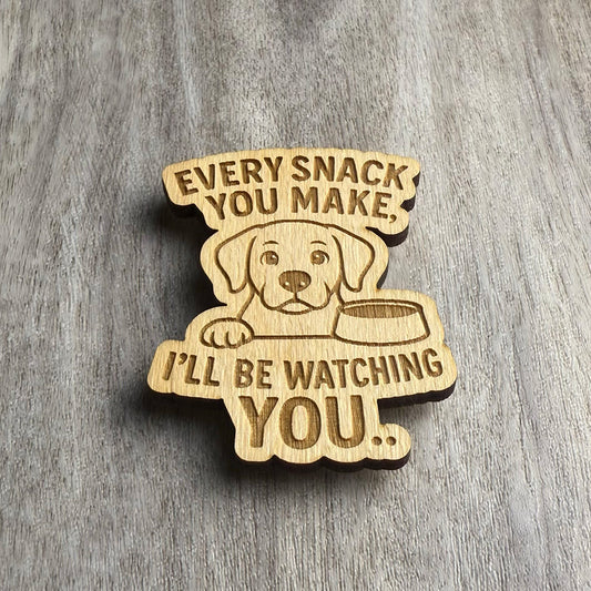 Front-facing view of a laser-cut wooden magnet with the engraved phrase Every Snack You Make, I’ll Be Watching You, including a dog illustration with a bowl, displayed on a gray wood background.