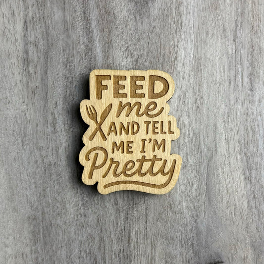 Laser-cut wooden magnet engraved with the phrase Feed Me and Tell Me I’m Pretty, featuring a fork and knife crossed in the center, styled with mixed fonts and placed on a gray wood background.
