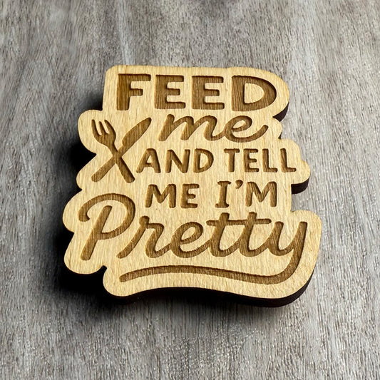 Angled view of a laser-cut wooden magnet showing the engraved phrase Feed Me and Tell Me I’m Pretty with a crossed fork and knife design, highlighting engraving depth and wood thickness on a gray surface.