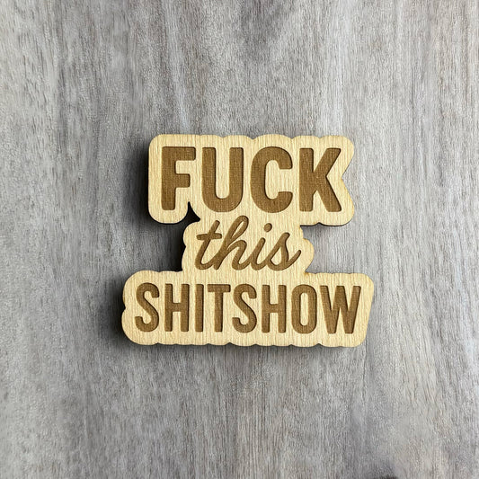 Laser-cut wooden magnet engraved with the phrase Fuck This Shitshow in a mix of bold and script fonts, placed on a gray wood background.