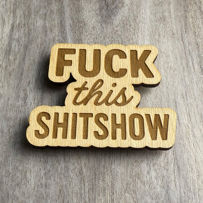 Angled view of a laser-cut wooden magnet displaying the phrase Fuck This Shitshow, showcasing the engraved detail and wood depth against a gray wood surface.