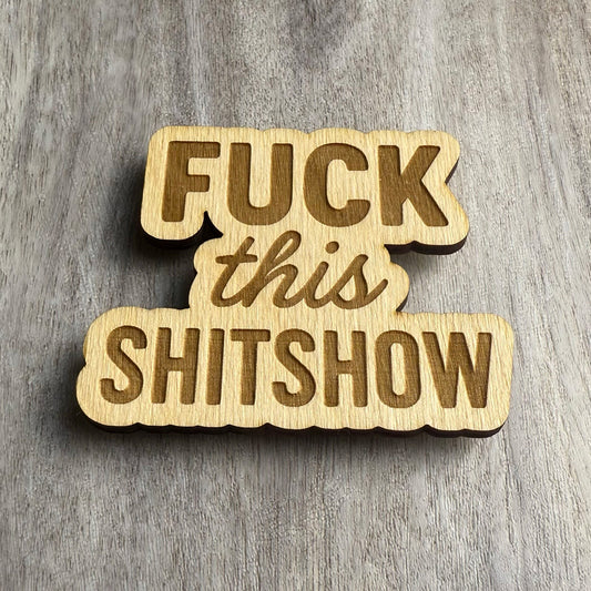 Angled view of a laser-cut wooden magnet displaying the phrase Fuck This Shitshow, showcasing the engraved detail and wood depth against a gray wood surface.