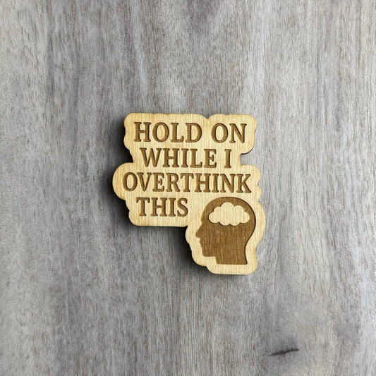 Laser-cut wooden magnet engraved with the phrase Hold On While I Overthink This, accompanied by a silhouette of a human head with a brain-shaped cloud inside, set on a gray wood background.