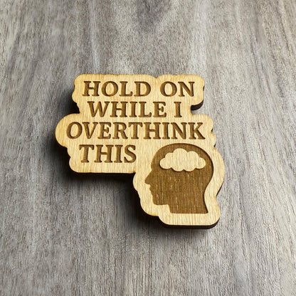 Angled view of a laser-cut wooden magnet featuring the phrase Hold On While I Overthink This and a head silhouette with a thought cloud, showing engraving depth and wooden thickness on a gray surface.