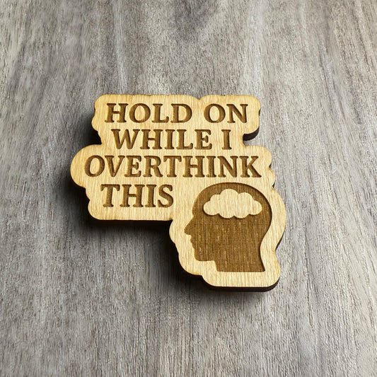 Angled view of a laser-cut wooden magnet featuring the phrase Hold On While I Overthink This and a head silhouette with a thought cloud, showing engraving depth and wooden thickness on a gray surface.