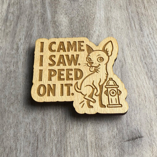 Angled view of a laser-cut wooden magnet with the phrase I Came I Saw I Peed On It and an illustration of a mischievous chihuahua peeing on a fire hydrant, showcasing engraving depth and material thickness on a gray surface.
