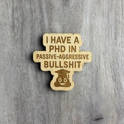 Laser-cut wooden magnet engraved with the phrase I Have a PhD in Passive-Aggressive Bullshit, featuring a smiling poop emoji wearing a graduation cap at the bottom, displayed on a gray wood background.