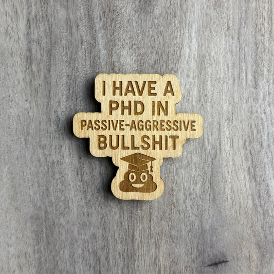 Laser-cut wooden magnet engraved with the phrase I Have a PhD in Passive-Aggressive Bullshit, featuring a smiling poop emoji wearing a graduation cap at the bottom, displayed on a gray wood background.