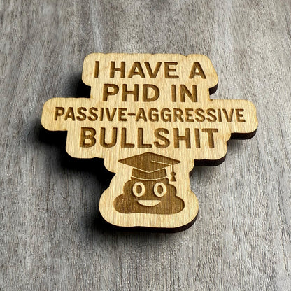 Angled view of a laser-cut wooden magnet showing the phrase I Have a PhD in Passive-Aggressive Bullshit with a poop emoji in a graduation cap, highlighting the engraving depth and wood layering on a gray surface.