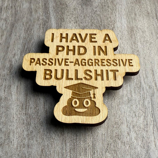 Angled view of a laser-cut wooden magnet showing the phrase I Have a PhD in Passive-Aggressive Bullshit with a poop emoji in a graduation cap, highlighting the engraving depth and wood layering on a gray surface.