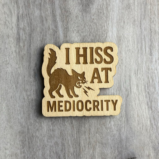 Laser-cut wooden magnet engraved with the phrase I Hiss at Mediocrity, featuring an angry, arched-back cat hissing with lightning bolts coming from its mouth, set against a gray wood background.