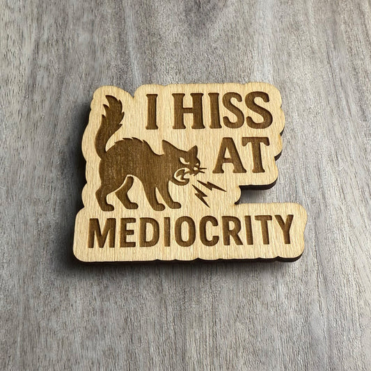 Angled view of a laser-cut wooden magnet showing the phrase I Hiss at Mediocrity and a fierce hissing cat illustration, highlighting engraving depth and wood thickness on a gray surface.