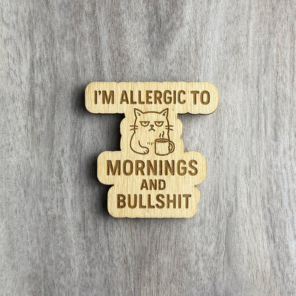 Laser-engraved wooden magnet featuring the phrase “I’M ALLERGIC TO MORNINGS AND BULLSHIT” in bold lettering. At the center is a grumpy-looking cat holding a steaming coffee mug, engraved in line art. The magnet has a custom cutout shape following the design elements.