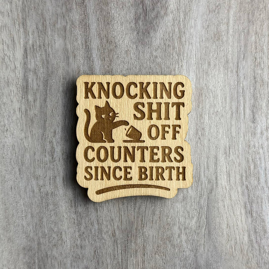 Laser-engraved wooden magnet with the phrase “KNOCKING SHIT OFF COUNTERS SINCE BIRTH.” The design features a mischievous cat swatting a cup off a surface, engraved in silhouette style. The magnet has a rounded rectangular shape with a slightly wavy edge.