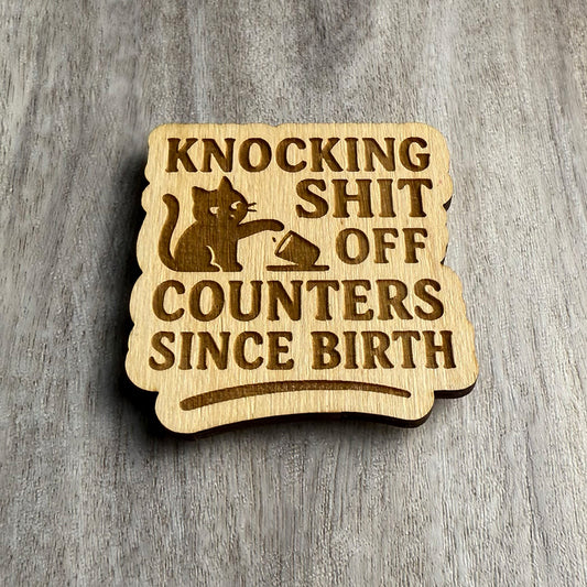 Angled view of the same engraved wooden magnet displaying the text “KNOCKING SHIT OFF COUNTERS SINCE BIRTH,” along with an engraving of a smug cat pawing a cup off an edge. The depth of the engraving and texture of the wood are highlighted by the lighting.