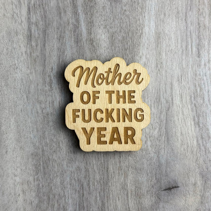 Laser-engraved wooden magnet with the phrase “Mother OF THE FUCKING YEAR.” The word “Mother” is in a cursive script, while the rest is in bold block letters. The magnet has a cloud-like, organic border shape that wraps around the stacked text.