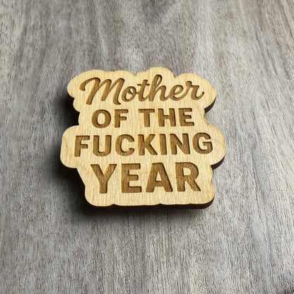 Angled view of the same engraved wooden magnet reading “Mother OF THE FUCKING YEAR,” combining elegant script and bold font styles. The lighting emphasizes the engraved depth and rounded edges of the custom cut shape.