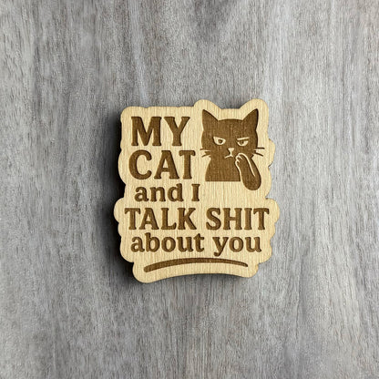 Laser-engraved wooden magnet with the phrase “MY CAT and I TALK SHIT about you.” The design includes a smug-looking cat with its paw to its mouth in a whispering pose. The magnet has a rounded rectangular shape with soft wave-like edges.