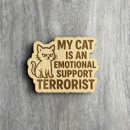 Laser-engraved wooden magnet with the phrase “MY CAT IS AN EMOTIONAL SUPPORT TERRORIST” in bold uppercase letters. A grumpy, evil-eyed cat illustration sits on the left side. The magnet has a rounded, irregular shape that follows the layout of the design.